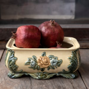 Peut inclure: Un bol décoratif en céramique avec une base crème et des motifs floraux verts et dorés ornés. Le bol contient trois grenades mûres à la peau rouge foncé. Le bol repose sur une surface en bois vieilli.