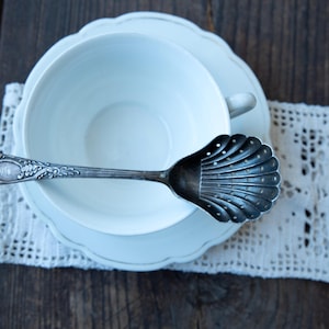 May include: A white teacup and saucer with a decorative silver spoon. The spoon has a shell-shaped bowl and ornate handle. The cup and saucer sit on a white lace doily, all on a dark wooden surface.