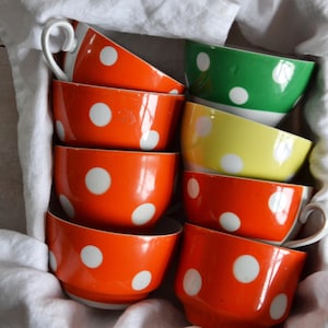 May include: A collection of brightly coloured ceramic teacups with white polka dots. The cups are in shades of orange, green, and yellow, and are arranged in a white cloth. The cups have white handles.