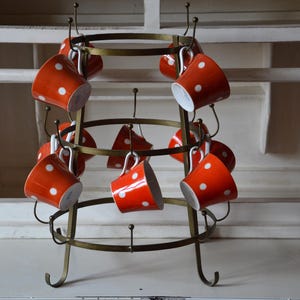 Può includere: Un portabicchieri vintage in ottone con dieci tazze in ceramica rossa. Ogni tazza presenta un motivo a pois bianchi. Il portabicchieri ha un design circolare con ganci curvi per le tazze.