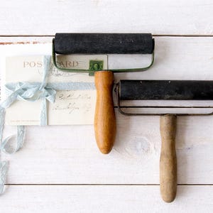 Puede incluir: Dos rodillos de impresión vintage con rodillos negros y mangos de madera sobre una superficie de madera blanca. Un montón de postales atadas con una cinta azul claro está en el fondo. La palabra "POSTCARD" es visible.