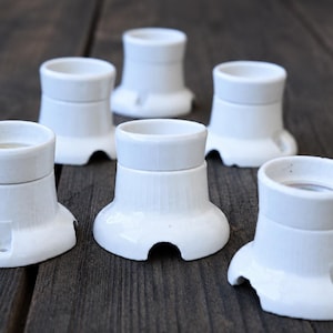 May include: A set of five white ceramic light bulb sockets with a flared base and a cylindrical top. Each socket features a smooth, glossy finish and small mounting holes at the base, designed for surface installation on a wall or ceiling.