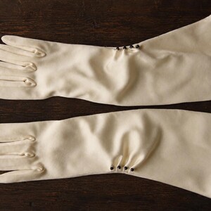 Peut inclure: Une paire de gants mi-longs blanc cassé en tissu lisse. Chaque gant présente une série de quatre petits ornements sphériques foncés placés le long de la zone extérieure du poignet, créant des plis subtils dans le matériau du tissu.