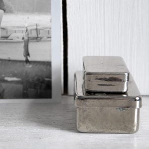 May include: Two rectangular silver-toned metal tins stacked on top of each other. The smaller tin rests on the lid of the larger tin. Both containers have a smooth, reflective surface and rounded corners, positioned next to a black and white photograph.