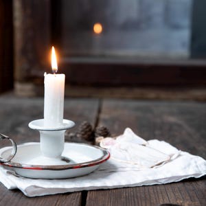 Può includere: Una candela bianca accesa in un candeliere di smalto bianco con bordo rosso, su un panno bianco. La fiamma della candela brilla. La scena è ambientata su una superficie di legno scuro.