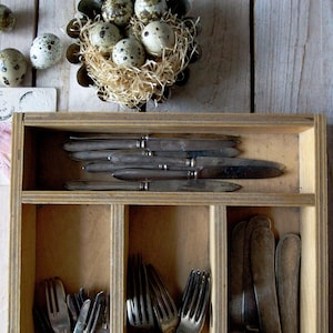 May include: A wooden cutlery organiser filled with vintage silverware, including knives and forks. A nest of speckled eggs sits above the organiser, with additional eggs scattered nearby. A card tied with pink ribbon is also present.