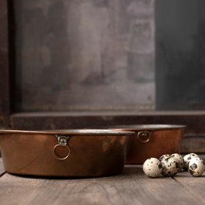 Op de afbeelding: Twee antieke koperen kommen met ringgrepen staan op een houten oppervlak, samen met verschillende gevlekte kwarteleieren. De kommen hebben een warme, roodbruine tint en de achtergrond omvat een rustiek raamkozijn.