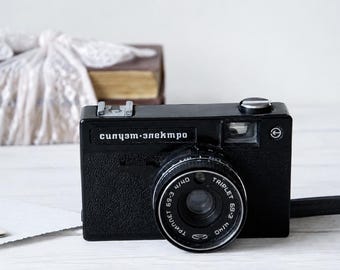 May include: A black vintage film camera with a textured body, featuring the Cyrillic text "силуэт-электро" on the front. The lens is labeled "Triplet 69-3 4/40" and includes a silver shutter button and a hot shoe mount on the top surface.