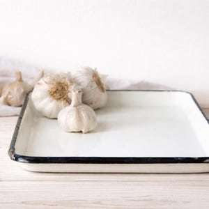 Può includere: Un vassoio rettangolare smaltato bianco con bordo nero, contenente diverse teste d'aglio. Il vassoio è appoggiato su una superficie di legno chiaro, con un panno bianco sullo sfondo. I bulbi d'aglio variano in dimensioni.