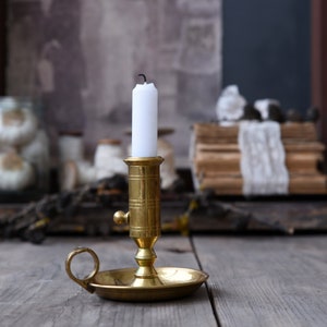 May include: A brass chamberstick candle holder with a circular base, a side handle loop, and a cylindrical socket featuring horizontal and vertical etched lines. A single white taper candle with a black wick is inserted into the socket.