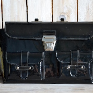 May include: A glossy black briefcase with a silver clasp and two front pockets. The vintage bag has a rectangular shape and a flap closure. The briefcase is made of a shiny material and is set against a light wooden background.