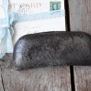 May include: A weathered, dark gray metal container with a textured surface. A vintage postcard tied with light blue lace ribbon is in the background. The postcard has the word "Brooklyn" and a stamp.