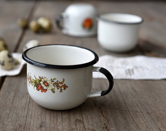 May include: A white enamel mug with a black rim and handle, featuring a floral decal with orange, yellow, and green flowers. The mug has a rounded body and a smooth finish. Additional matching mugs and small speckled eggs are visible in the background.