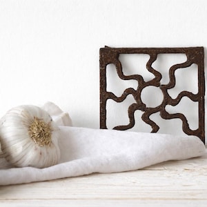 May include: A square, brown metal trivet with a sunburst pattern. Three garlic bulbs and a white cloth are in the foreground. The trivet is on a white wooden surface.