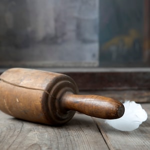 May include: A wooden rolling pin with a handle, lying on a wooden surface. The rolling pin is a light brown color and has a crack running across the top. A white feather duster is next to the rolling pin.