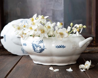 May include: A white ceramic serving dish with a blue heron design and floral accents, filled with white flowers. The dish has a lid and a handle. The flowers are white with yellow centers. The dish is on a dark wooden surface.