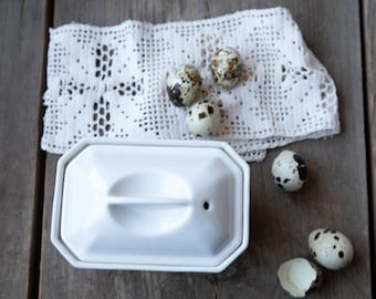 May include: A white ceramic butter dish with a lid, alongside several speckled quail eggs and a white lace doily. The dish has a rectangular shape with a handle on top. The eggs are small, with brown and black spots.
