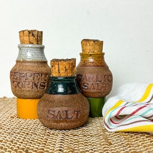 Pode incluir: Três potes de especiarias de cerâmica com rolhas de cortiça. Os potes estão rotulados como "Pepper Corns", "Salt" e "Garlic Salt". Os potes têm uma aparência rústica e artesanal com uma base marrom e detalhes coloridos. Uma toalha de cozinha listrada está no fundo.