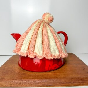 May include: A red teapot with a knitted pink and white striped tea cozy with a pom pom on top. The teapot is sitting on a wooden surface.