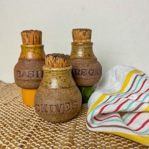 Pode incluir: Três pequenos potes de especiarias de cerâmica artesanais com rolhas de cortiça. Os potes estão rotulados como "BASIL", "OREGAN" e "CHIVES". Os potes são castanhos e amarelos e estão sobre uma esteira tecida com uma toalha listrada.