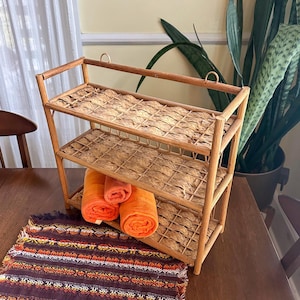 Op de afbeelding: Een drielaags, lichtbruin bamboe rek met geweven planken. Op de onderste plank liggen opgerolde oranje handdoeken. Onder het rek ligt een patroontextiel met oranje, bruine en witte ontwerpen.