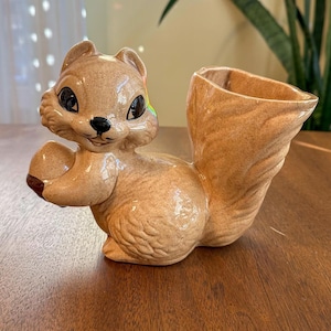 Könnte beinhalten: Ein keramischer Eichhörnchen-Pflanztopf in Braun- und Beigetönen. Das Eichhörnchen hält eine Nuss und hat eine große Öffnung auf dem Rücken für Pflanzen. Das Eichhörnchen hat schwarze Augen und eine glänzende Oberfläche. Ein dekorativer Artikel für die Inneneinrichtung.