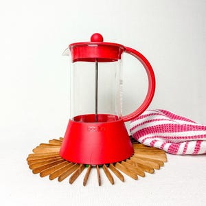 May include: A red Bodum French press coffee maker with a clear glass carafe. The coffee maker sits on a wooden trivet. A red and white striped towel is in the background.