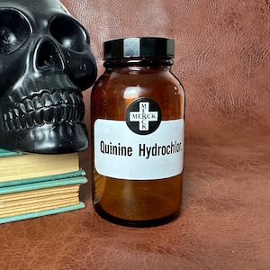 Könnte beinhalten: Eine braune Glasflasche mit einem schwarzen Schraubverschluss und einem weißen Etikett mit der Aufschrift "Quinine Hydrochlor.". Die Flasche hat ein schwarz-weißes Kreuz mit dem Wort "MERCK" im Kreuz. Die Flasche steht auf einer braunen Lederoberfläche.