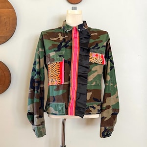 Puede incluir: Chaqueta con estampado de camuflaje con mangas largas, con una franja rosa en el centro y ribete de satén negro. La chaqueta tiene dos bolsillos con parches con estampado de tigre. La chaqueta se exhibe en un maniquí.