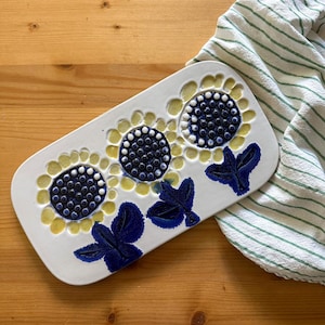 May include: A rectangular ceramic serving platter with a sunflower design. The platter features three sunflowers with yellow petals and dark blue centers. Blue leaves are painted at the base of the flowers. A green and white striped cloth is in the background.