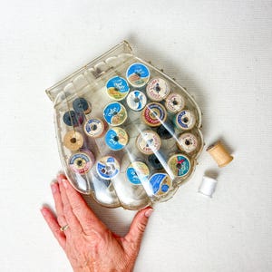 May include: A clear, seashell-shaped container filled with vintage spools of thread. The spools are various colors and sizes, with labels that read "Dual Duty" and "Clark's". A wooden spool and a white spool are next to the container.