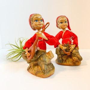 May include: Two vintage figurines of elves, each with a wooden face, blue eyes, and a red outfit. One elf holds a lute, the other a harp. They sit on a brown base. A small air plant is in the background.