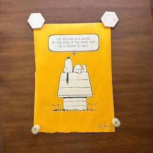 May include: A bright yellow rectangular poster featuring a black and white line drawing of Snoopy lying on top of a doghouse. A speech bubble above contains the text: LET ME LIVE IN A HOUSE BY THE SIDE OF THE ROAD AND BE A FRIEND TO MAN. The name Schulz is printed in the bottom right corner.