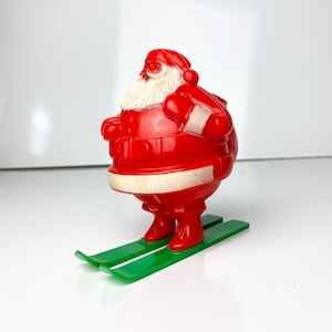May include: A vintage red Santa Claus figurine with a white beard and hat trim, standing on green skis. The figure has a white belt and is holding a sack. A festive Christmas decoration.