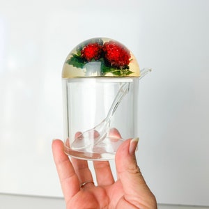 May include: A clear glass sugar dispenser with a dome lid featuring a red and green pressed flower design. The dispenser includes a small spoon.