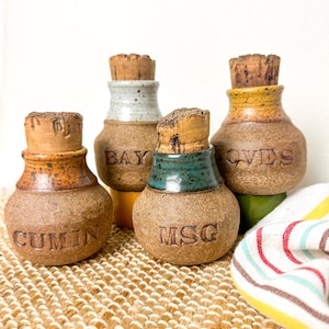 Pode incluir: Quatro potes de especiarias de cerâmica artesanais com rolhas de cortiça. Os potes estão rotulados com "CUMIN", "BAY", "MSG" e "CLOVES". Cada pote tem um esquema de cores único, com detalhes em castanho, amarelo e turquesa. Os potes estão dispostos sobre uma esteira tecida.