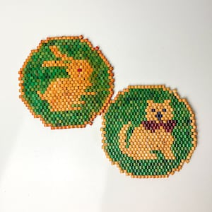May include: Two round wooden coasters with green and orange designs. One coaster features a rabbit and the other features a dog.