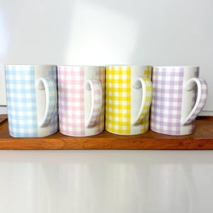 Könnte beinhalten: Vier Keramikbecher mit Gingham-Mustern in Blau, Rosa, Gelb und Lila. Die Becher stehen auf einer Holzoberfläche.