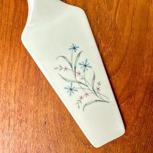 May include: White ceramic cake server with a floral design featuring blue and pink flowers. The handle is white and the blade is slightly curved.