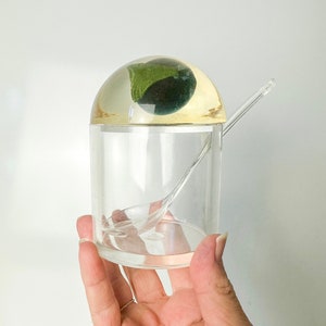 Può includere: Un contenitore cilindrico trasparente con un coperchio arrotondato tinto di giallo. Il coperchio racchiude un oggetto di forma organica verde scuro. Un cucchiaio trasparente è all'interno del contenitore. Il contenitore è tenuto in una mano.