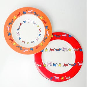 Könnte beinhalten: Zwei Keramikplatten mit weißen Zentren und farbigen Rändern. Eine Platte hat einen orangefarbenen Rand, die andere einen roten Rand. Beide Platten zeigen ein sich wiederholendes Muster von Cartoon-Katzen in verschiedenen Farben.