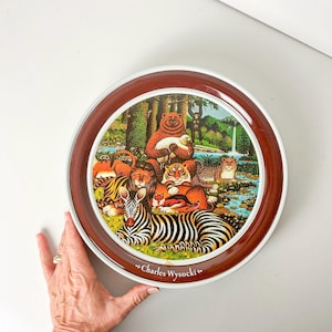 Könnte beinhalten: Ein runder, braun-weißer Metallteller mit einer farbenfrohen Illustration von Tieren in einer Waldlandschaft. Auf dem Teller sind ein Bär, der ein Lamm hält, ein Zebra, ein Löwe, ein Tiger und ein Leopard zu sehen. Der Text "~Charles Wysocki~" ist auf der Unterseite des Tellers aufgedruckt.
