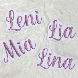 Könnte beinhalten: Vier gestickte Namensaufnäher: Leni, Lia, Mia und Lina. Jeder Name ist in einer mauvefarbenen Schreibschrift auf einen weißen, wolkenförmigen Aufnäher gestickt. Die Aufnäher liegen auf einer weißen, strukturierten Oberfläche.