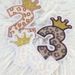 Könnte beinhalten: Gestickte Stoffzahlen 1, 2 und 3 mit goldenen Glitzerkronen. Die Zahlen haben ein Leopardenmuster in Braun- und Beigetönen. Die Zahlen sind weiß umrandet.