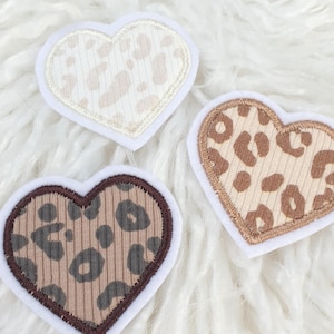 Könnte beinhalten: Drei herzförmige Aufnäher mit Leopardenmuster. Jeder Aufnäher hat einen weißen Rand und ein anderes Farbschema: Creme, Braun und Beige. Die Aufnäher sind auf einer flauschigen weißen Oberfläche angeordnet.