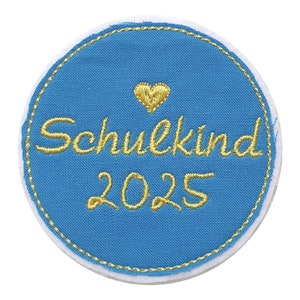 Könnte beinhalten: Ein runder, blauer Aufnäher mit dem goldfarbenen Schriftzug "Schulkind 2025" und einem goldenen Herz. Der Aufnäher hat einen weißen Rand und goldfarbene Nähte.