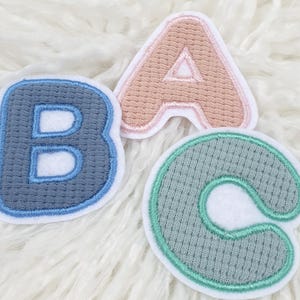 Könnte beinhalten: Drei gestickte Stoffaufnäher mit den Buchstaben "B A C". Der Buchstabe "B" ist blau mit einer dunkelblauen Umrandung. Der Buchstabe "A" ist pfirsichfarben mit einer rosa Umrandung. Der Buchstabe "C" ist grün mit einer türkisfarbenen Umrandung.