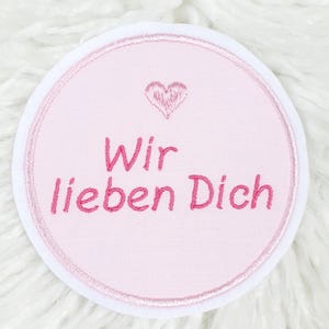 Könnte beinhalten: Ein runder, rosa bestickter Aufnäher mit dem deutschen Text "Wir lieben Dich" in rosa Schrift, was "Wir lieben dich" bedeutet. Ein kleines rosa Herz ist über dem Text gestickt. Der Aufnäher hat einen weißen Rand und ist vor einem flauschigen weißen Hintergrund platziert.