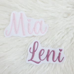 Könnte beinhalten: Zwei bestickte Namensaufnäher. Einer zeigt "Mia" in hellrosa Schrift, der andere "Leni" in dunklerer rosa Schrift. Beide Aufnäher sind auf weißem Filz.