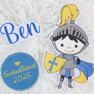 Könnte beinhalten: Gestickte Aufnäher mit einem Cartoon-Ritter mit gelbem Schild und blauen Akzenten, einem blauen Aufnäher mit dem Namen "Ben" und einem runden Aufnäher mit "Schulkind 2025" in goldfarbenen Buchstaben. Auf weißem, flauschigem Untergrund.